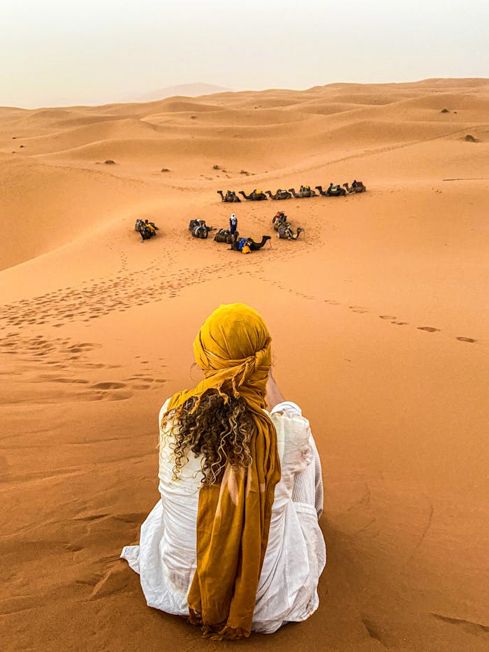 A woman gazes over a camel caravan in the Moroccan desert at sunset, serene and picturesque.